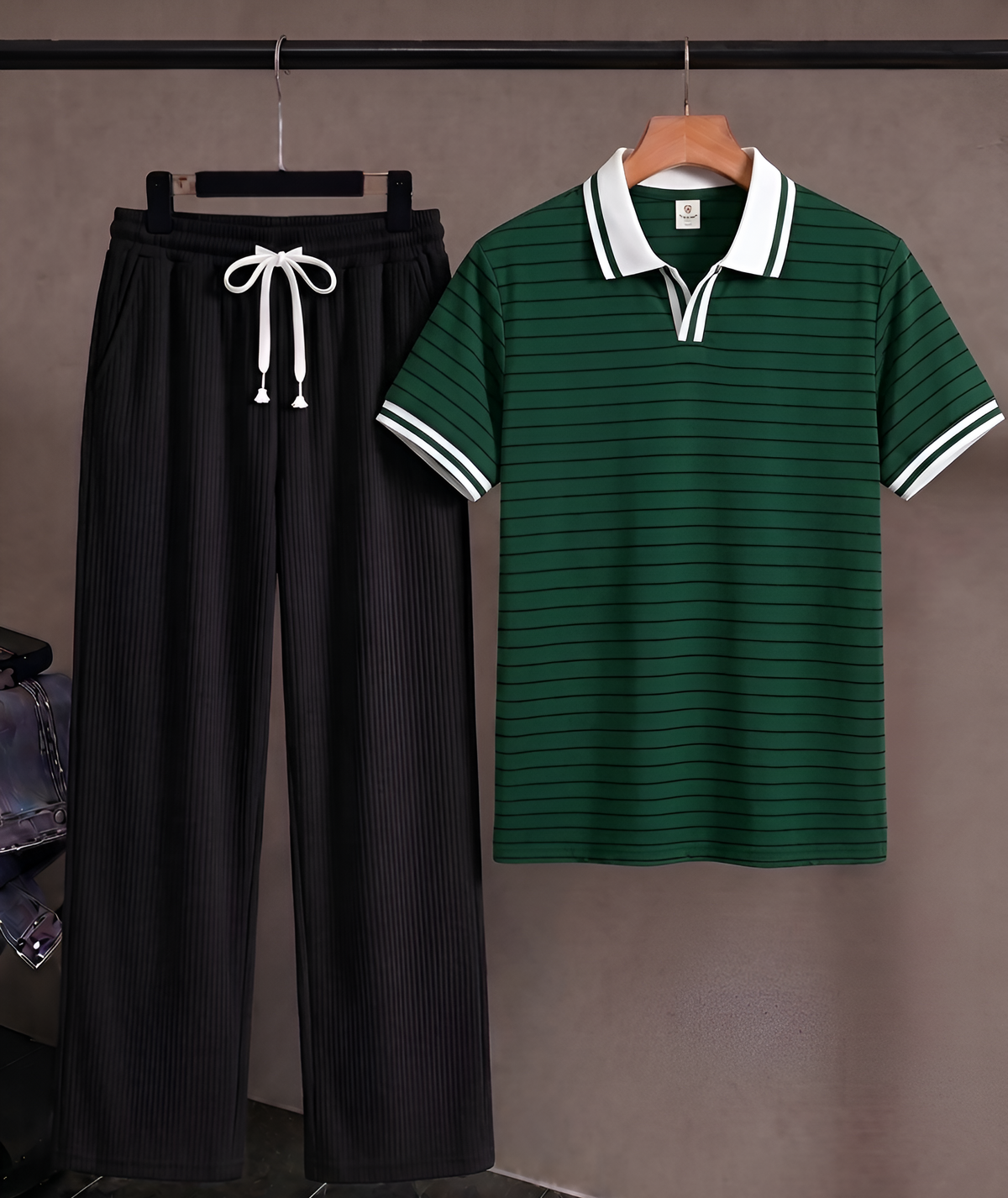 A two-piece clothing set hanging on a black rack against a taupe wall, featuring a dark green short-sleeve polo shirt with white striped trim and matching black pleated drawstring trousers.